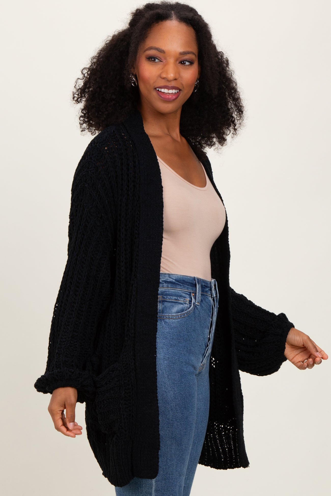 woman wearing a black oversized knit cardigan over a beige top and blue jeans, smiling and posing gently