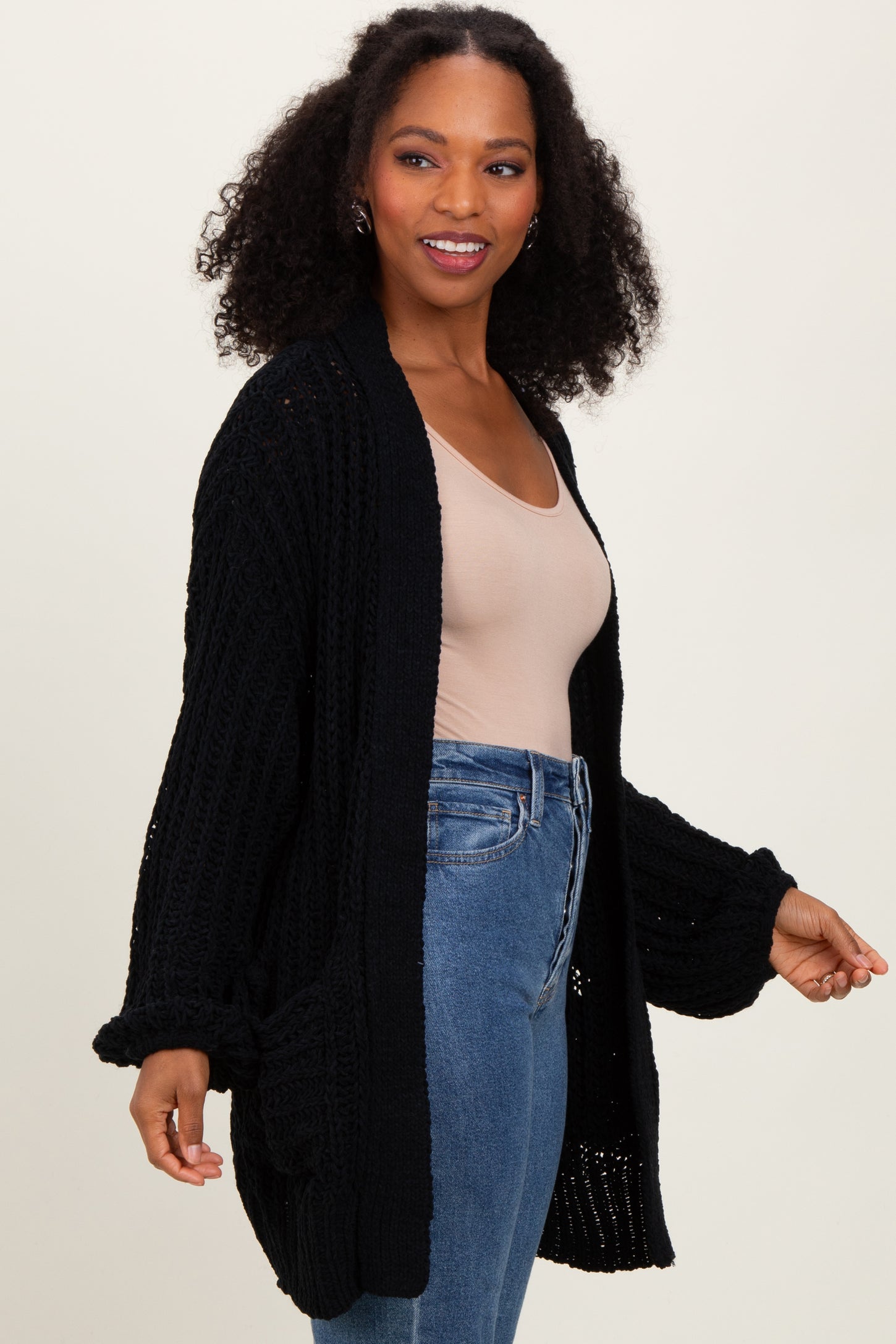woman wearing a black oversized knit cardigan over a beige top and blue jeans, smiling and posing gently