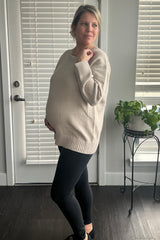 woman wearing a beige maternity sweater and black leggings, showing her baby bump while standing by a window