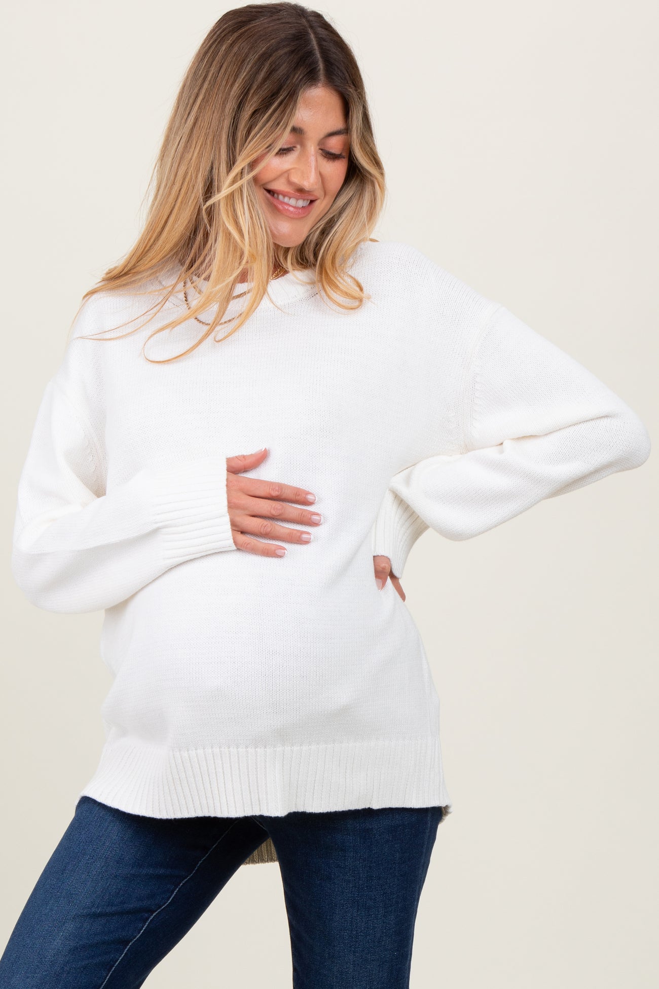 woman wearing a relaxed fit white maternity sweater and holding her baby bump, paired with denim jeans