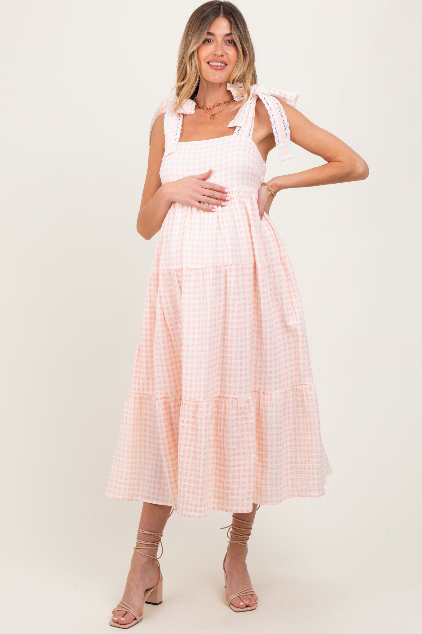 woman wearing a pink and white gingham maternity dress with tie straps and tiered skirt, holding her baby bump