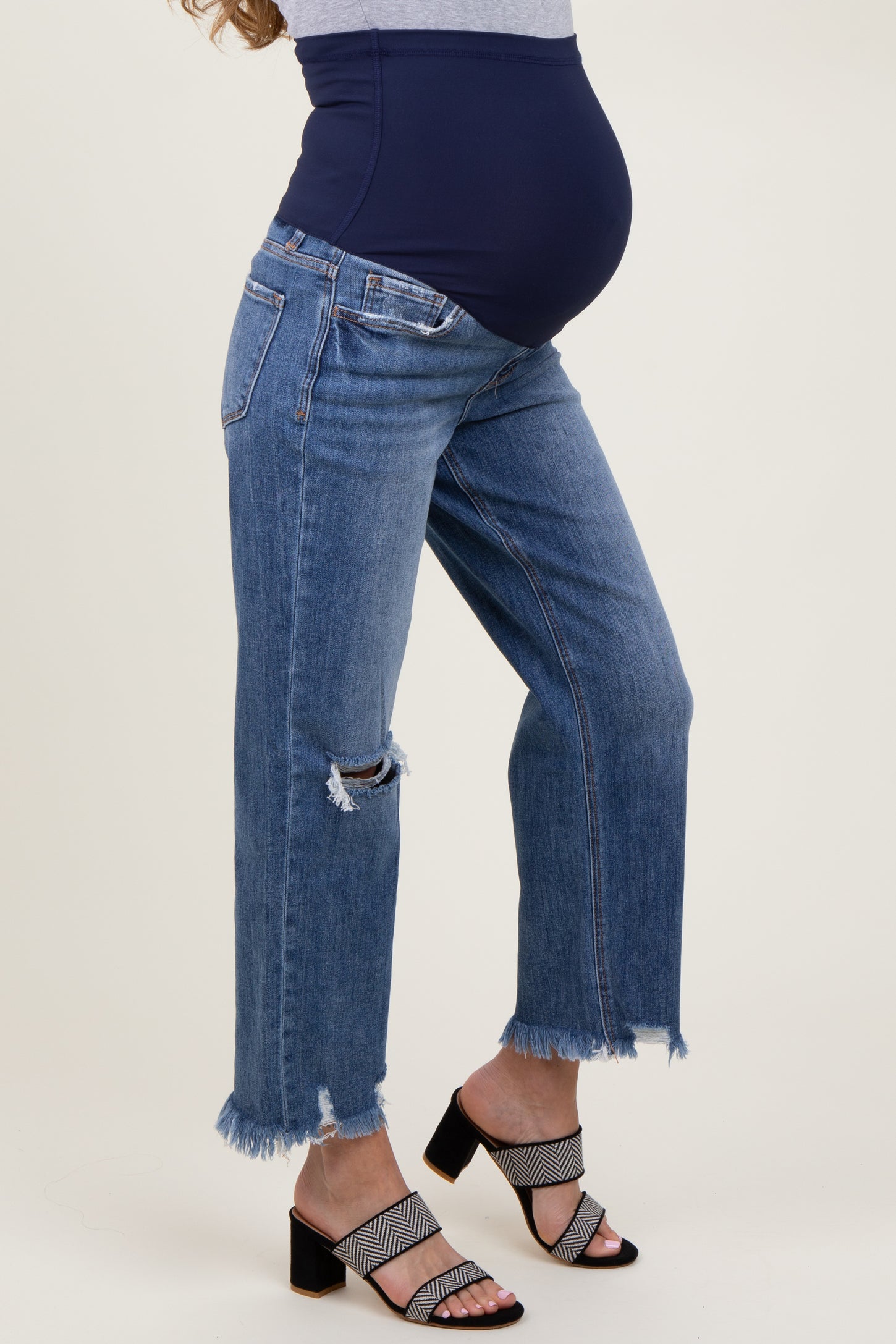woman wearing distressed blue maternity jeans with a dark blue over-the-bump panel and stylish black and white heeled sandals, showing baby bump