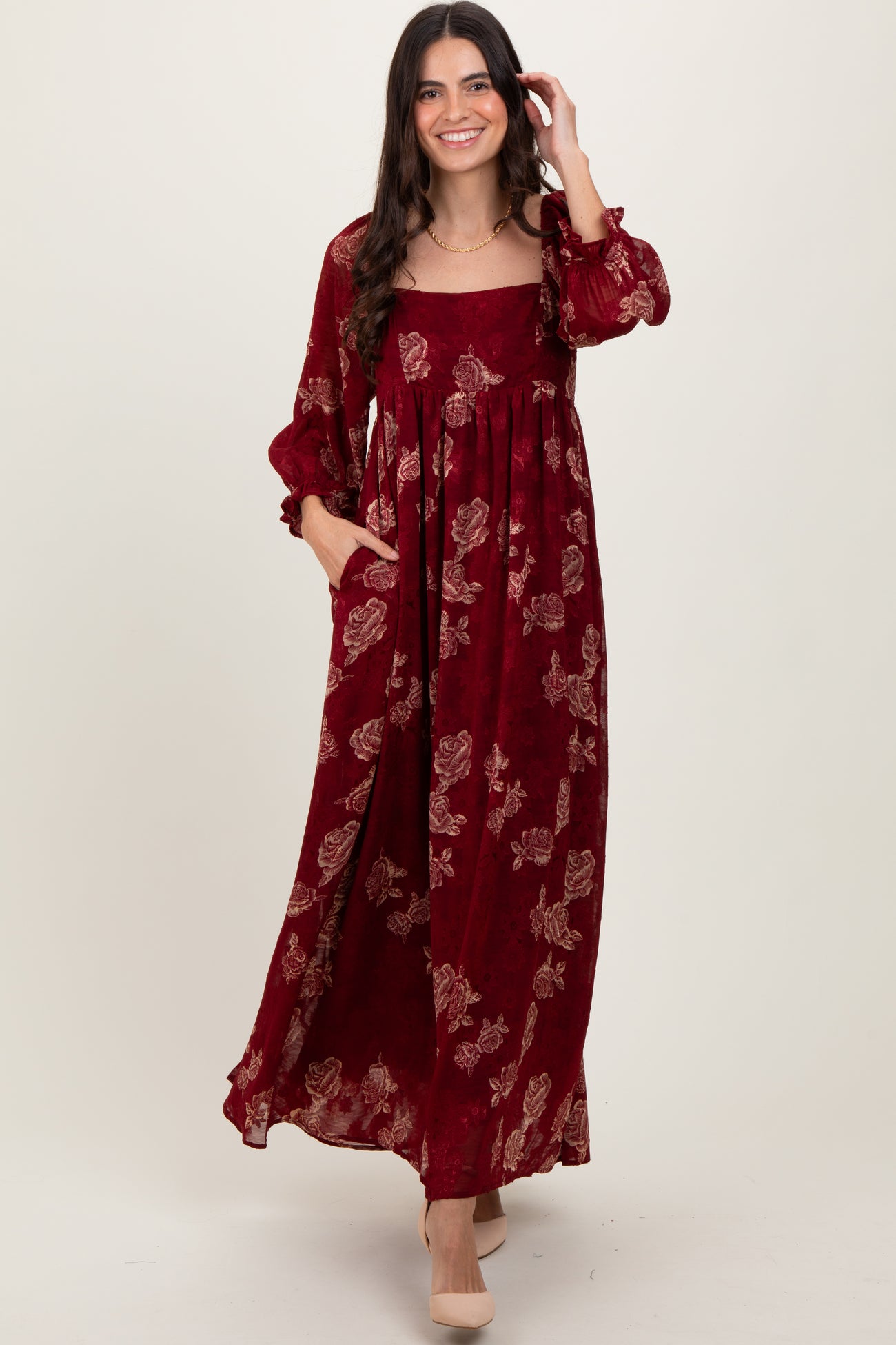 woman wearing a burgundy floral maxi dress with long sleeves and pockets, smiling at the camera