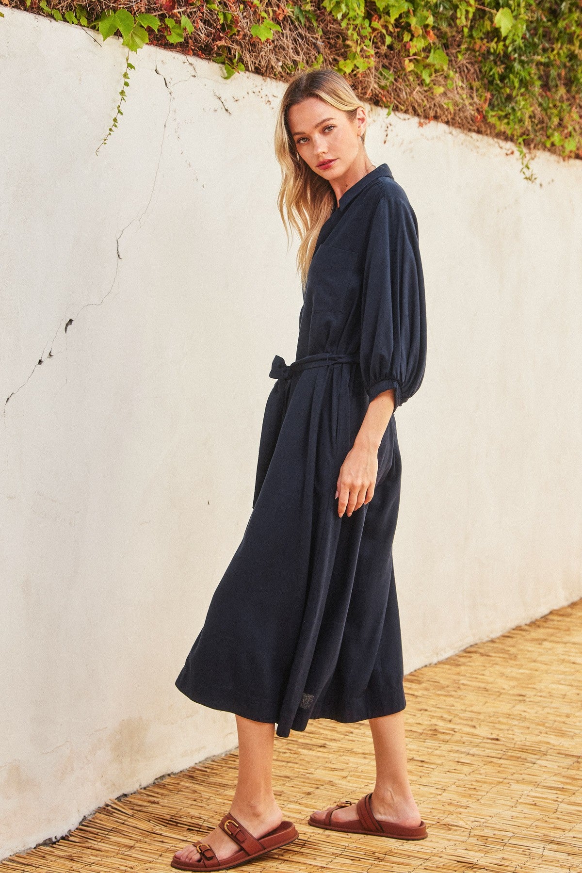 woman wearing a navy blue midi dress with balloon sleeves and a tie waist detail, standing against a textured wall