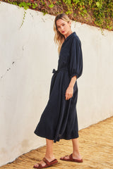 woman wearing a navy blue midi dress with balloon sleeves and a tie waist detail, standing against a textured wall
