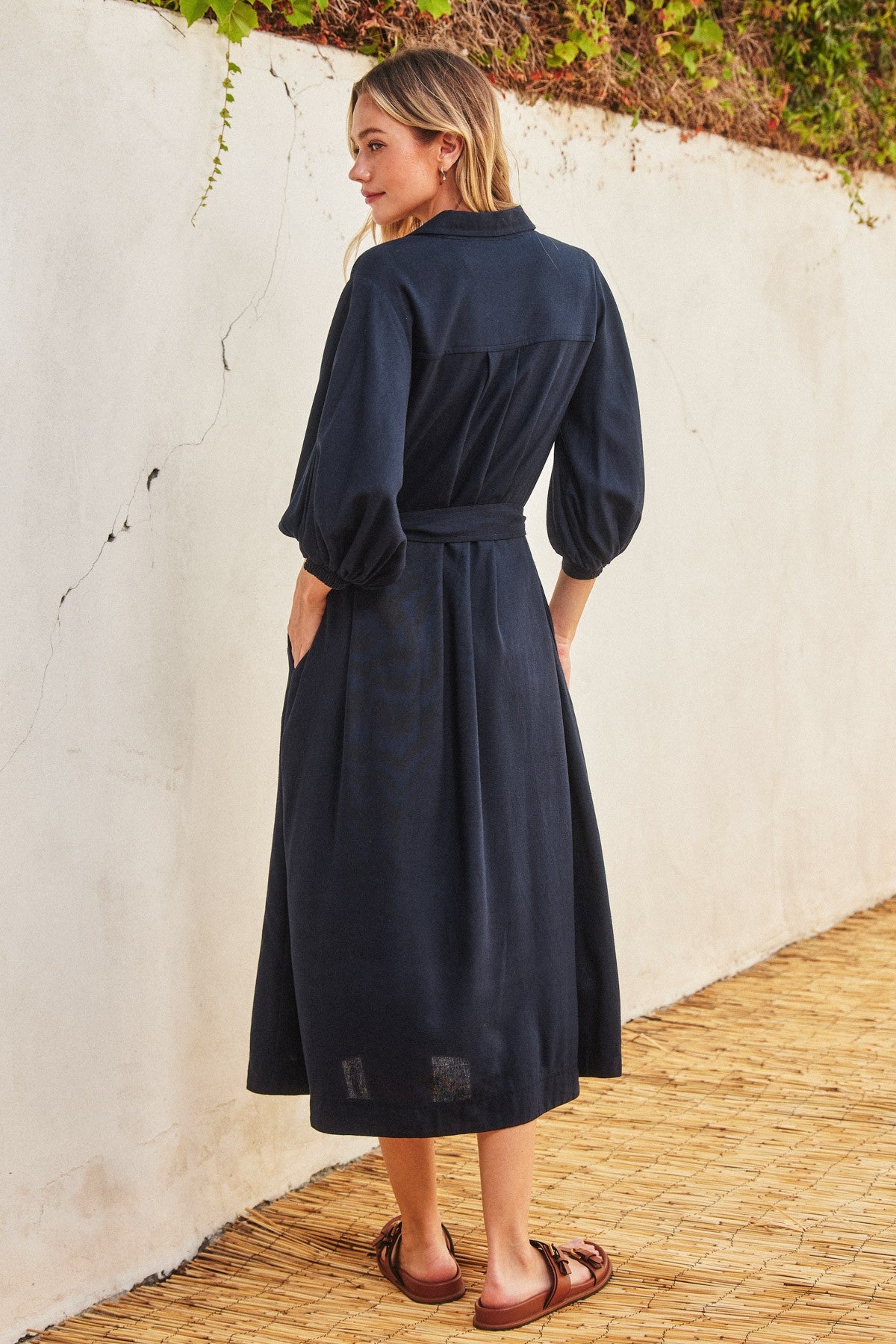 woman wearing a navy blue midi dress with puff sleeves and a belted waist, standing against a textured wall