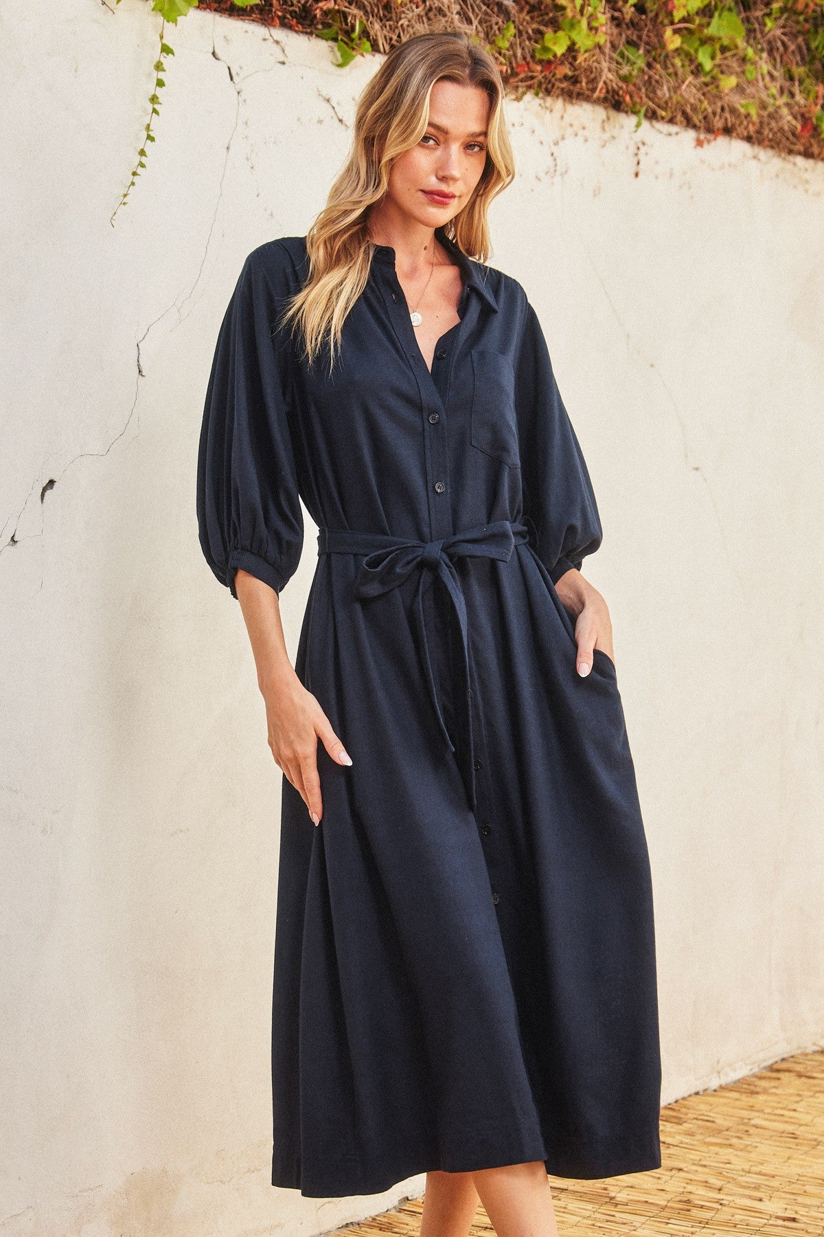 woman wearing a navy blue button-up midi dress with puff sleeves and a tied waist, standing against a textured wall