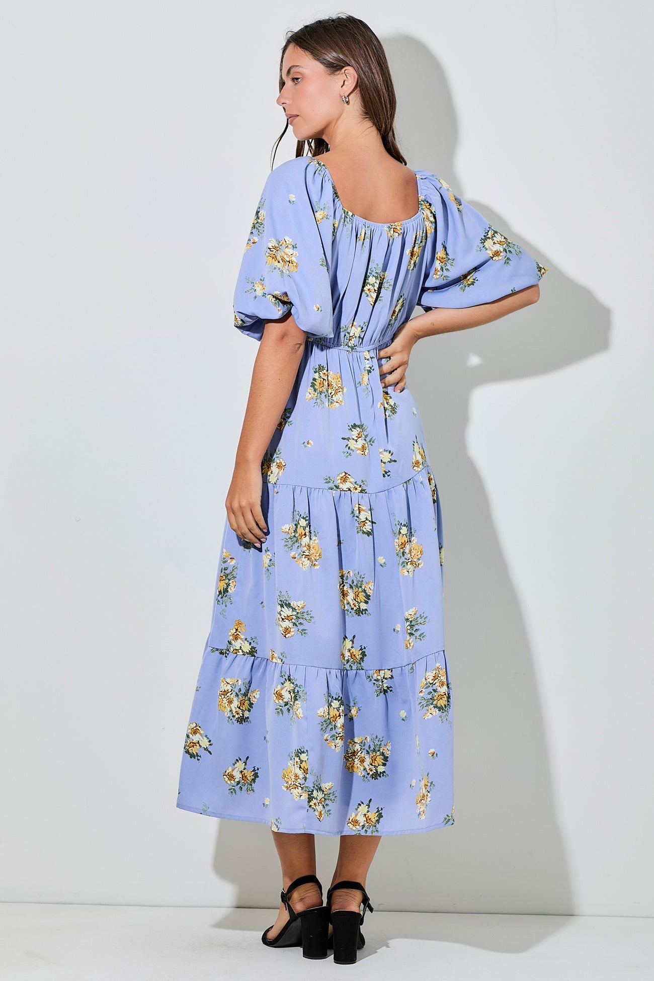 woman wearing a light blue floral maxi dress with puff sleeves and a tiered skirt, standing with her back to the camera