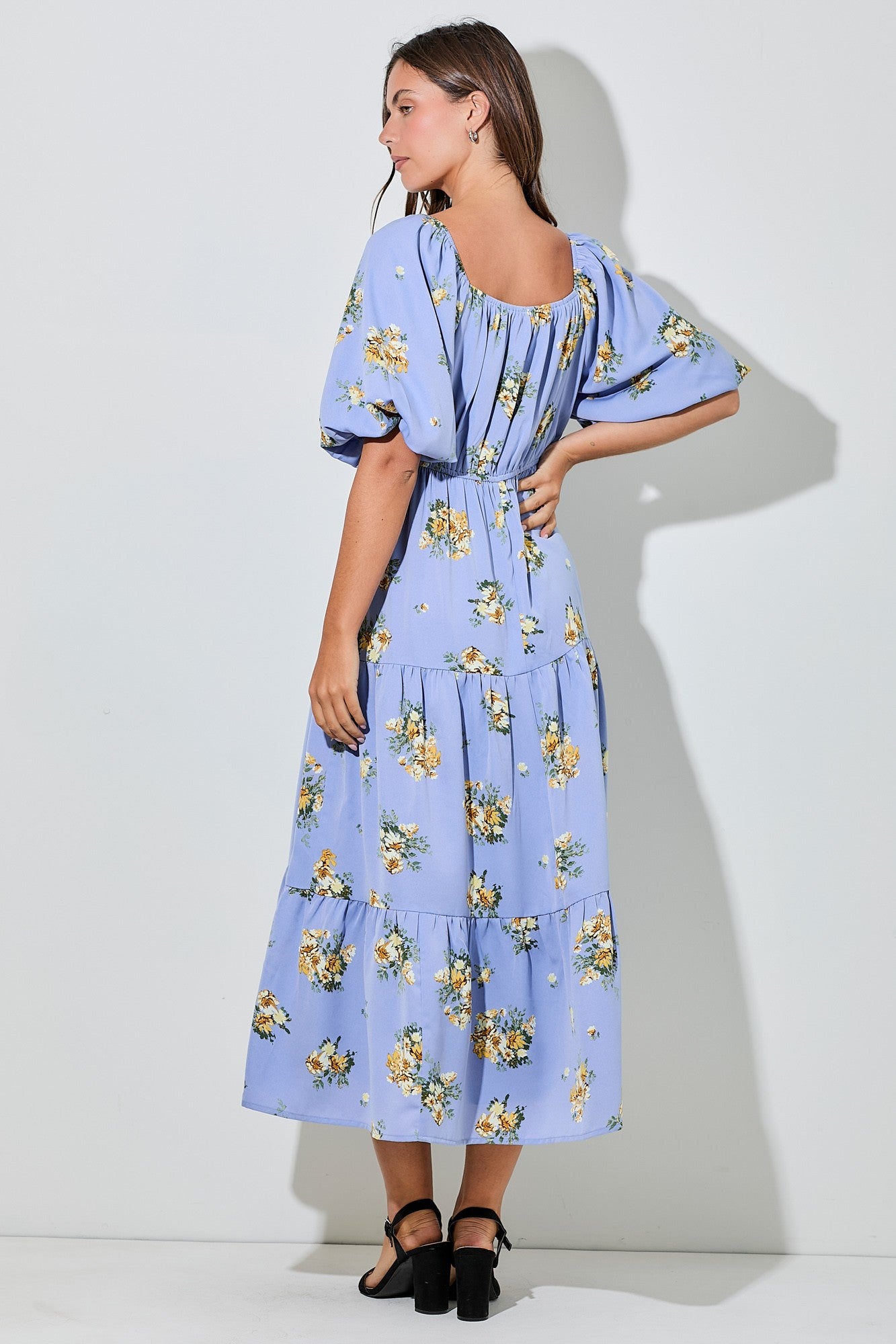 woman wearing a light blue floral maxi dress with puff sleeves and a tiered skirt, standing with her back to the camera