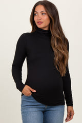 woman wearing a black long sleeve maternity turtleneck top and showing her baby bump while standing against a light background