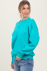 woman wearing a bright turquoise oversized sweatshirt and blue jeans, standing with a slight smile