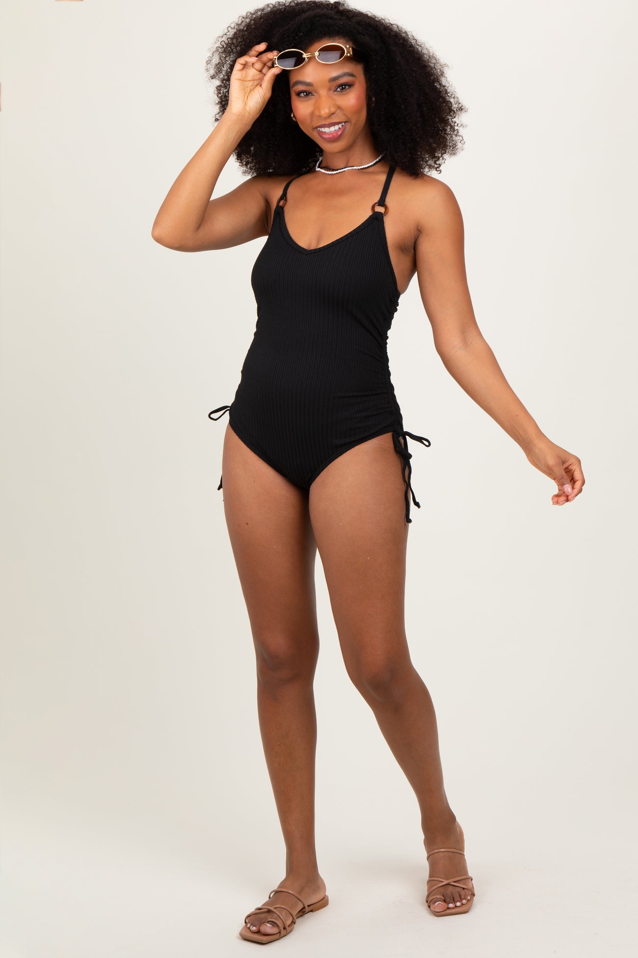 woman wearing a black ribbed one-piece swimsuit with adjustable side ties and holding sunglasses