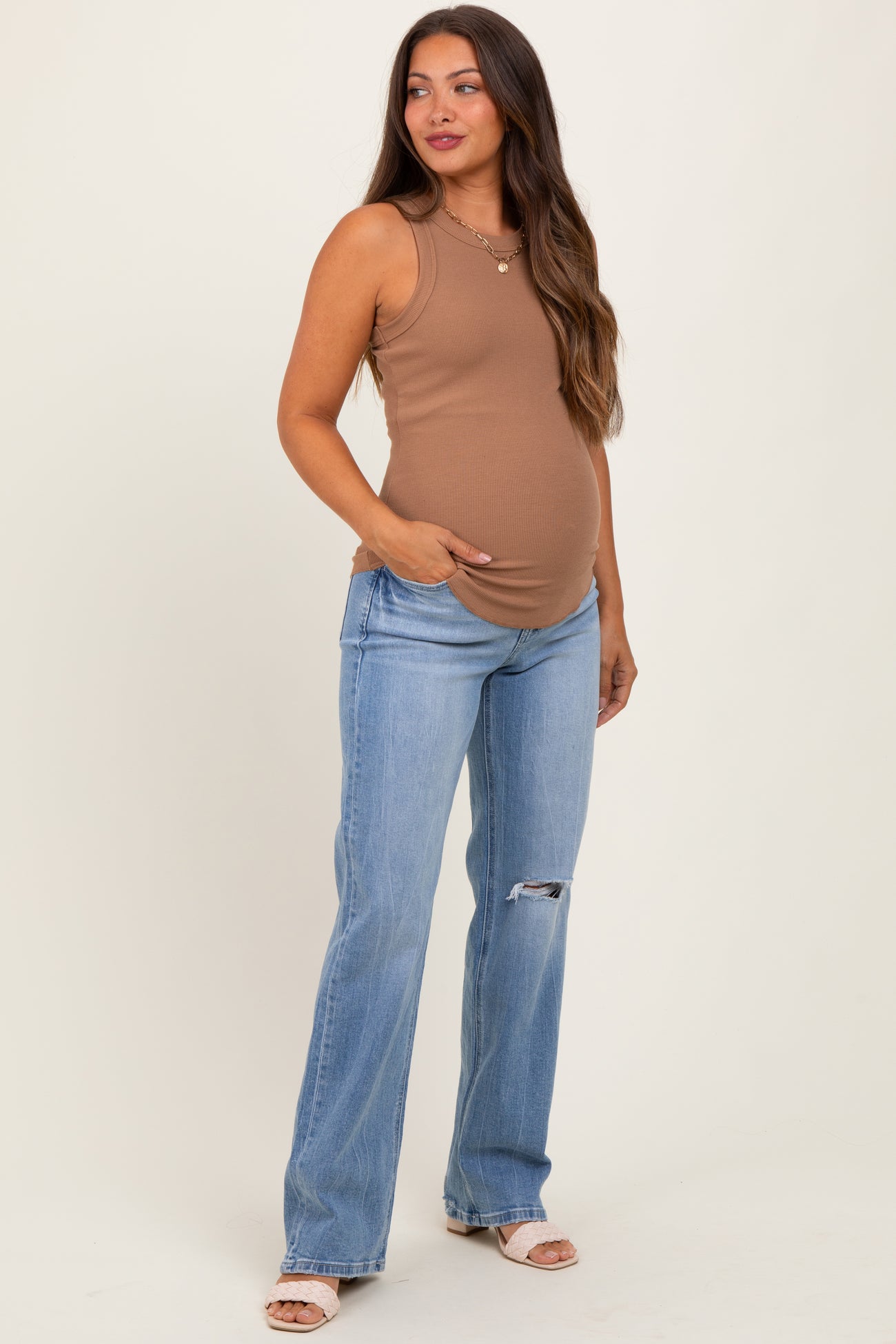woman wearing a brown sleeveless maternity tank top and blue distressed maternity jeans, showing her baby bump