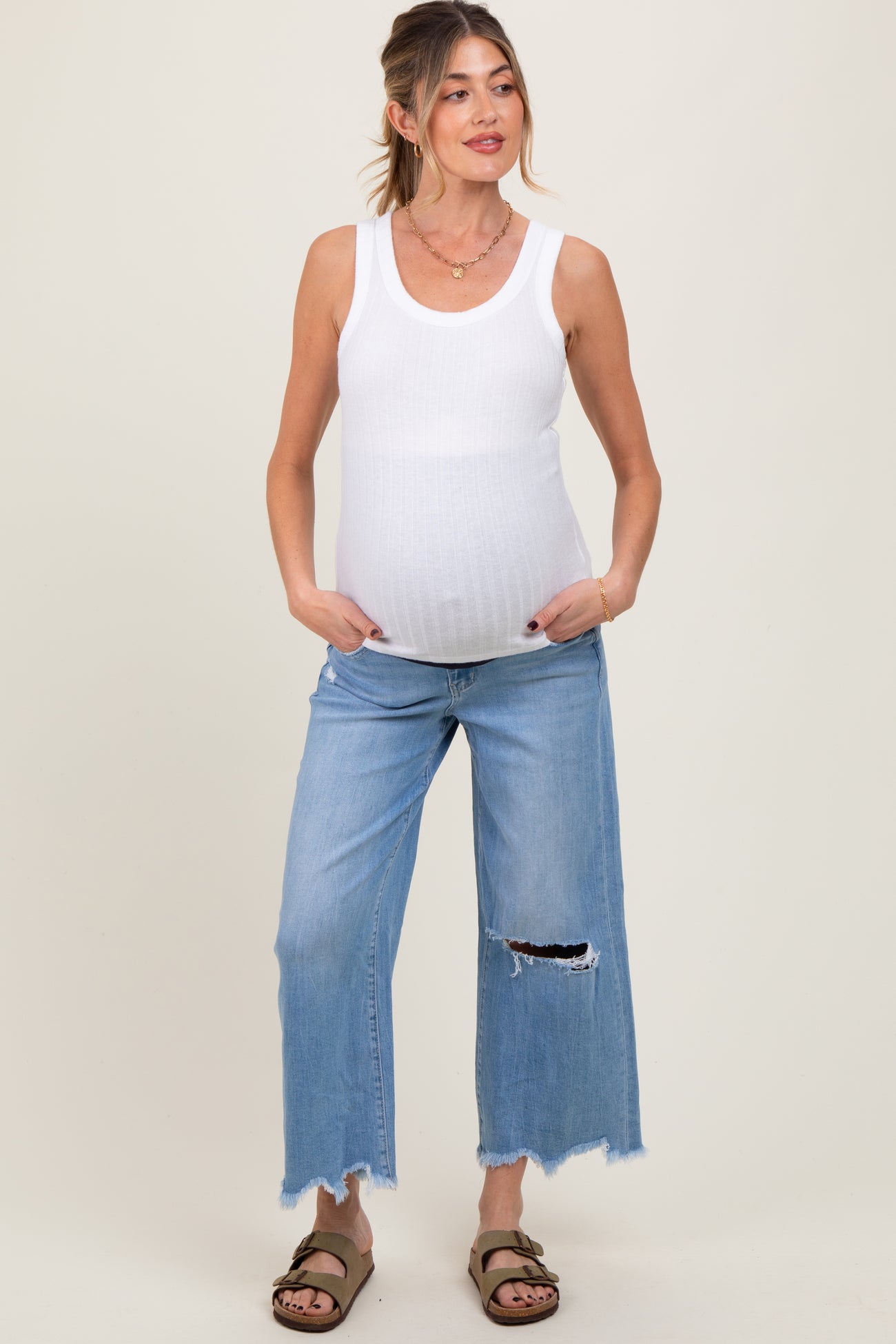woman wearing a white ribbed maternity tank top and distressed light blue maternity jeans with a visible baby bump