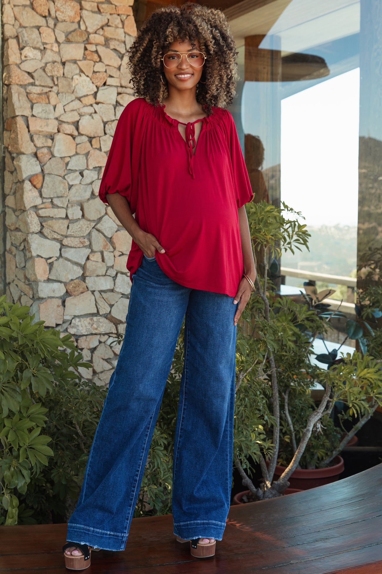 woman wearing a red maternity top with a tie neckline and blue wide-leg jeans, showing her baby bump