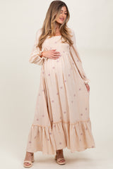 woman wearing a long sleeve beige maternity dress with floral embroidery and a ruffled hem, holding her baby bump