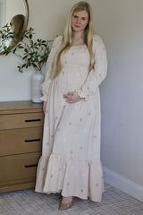 woman wearing a light pink maternity dress with floral embroidery and smocked bodice, holding her baby bump