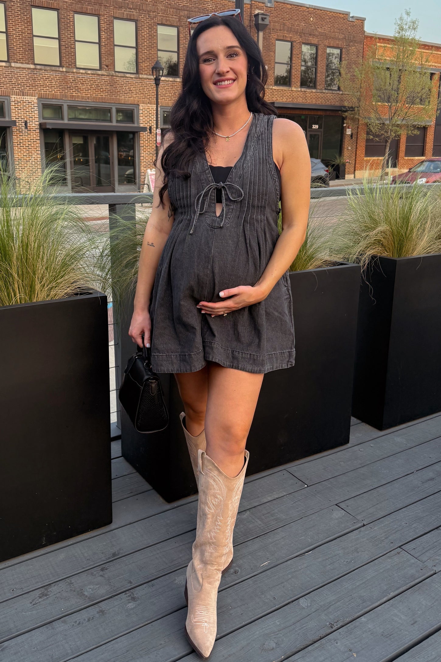 woman wearing a black maternity romper with tied front detail and posing with her baby bump in stylish knee-high boots