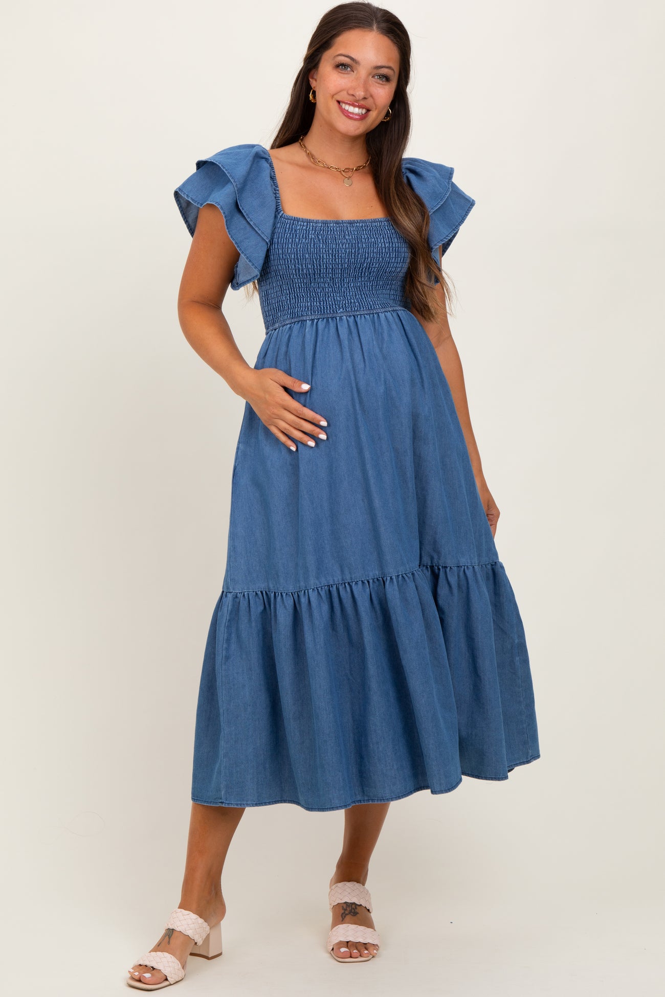 woman wearing a blue denim maternity dress with ruffled sleeves and a smocked bodice, holding her baby bump