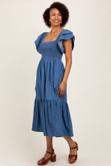 woman wearing a blue denim midi dress with flutter sleeves and a smocked bodice, paired with brown block-heeled sandals