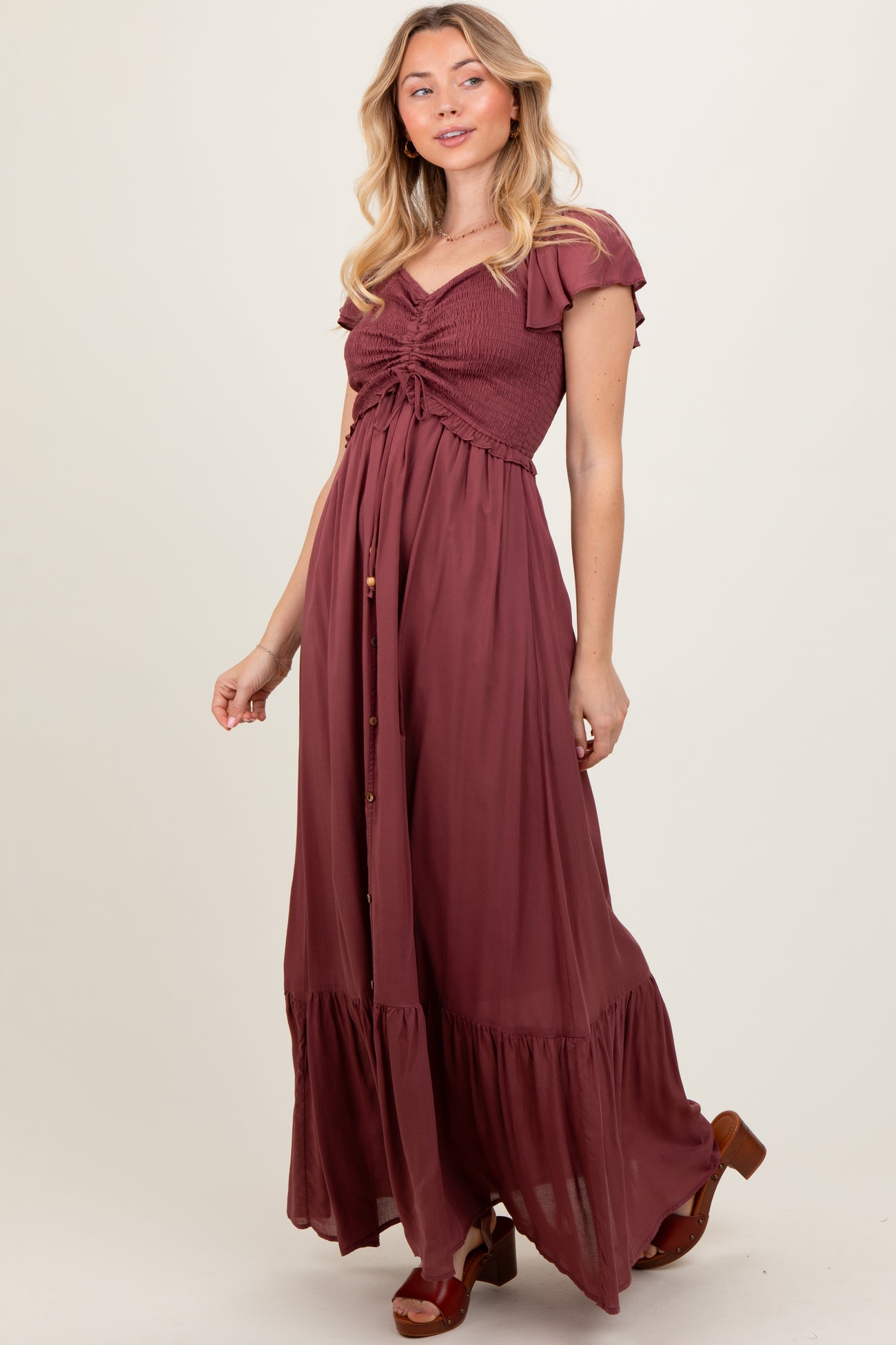 woman wearing a burgundy maxi dress with a smocked bodice, flutter sleeves, and button details, standing and smiling