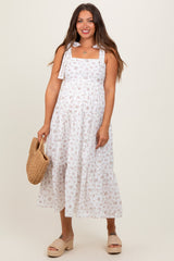 woman wearing a white floral maternity dress with tie straps and a flowy skirt, holding a woven bag and showing her baby bump