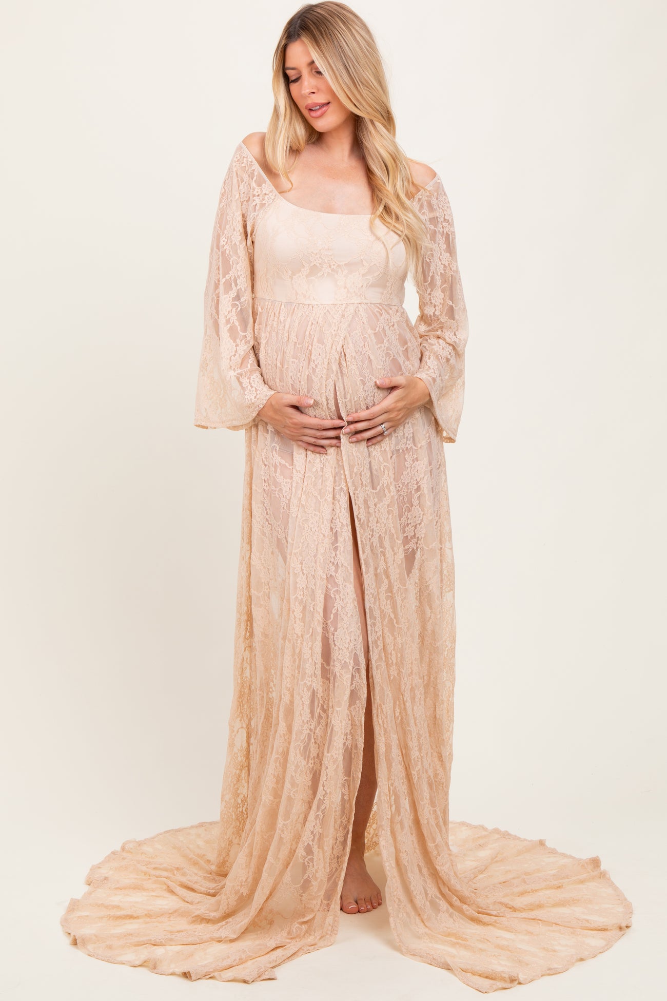 woman wearing a sheer beige lace maternity gown with long sleeves and a flowing train, holding her baby bump