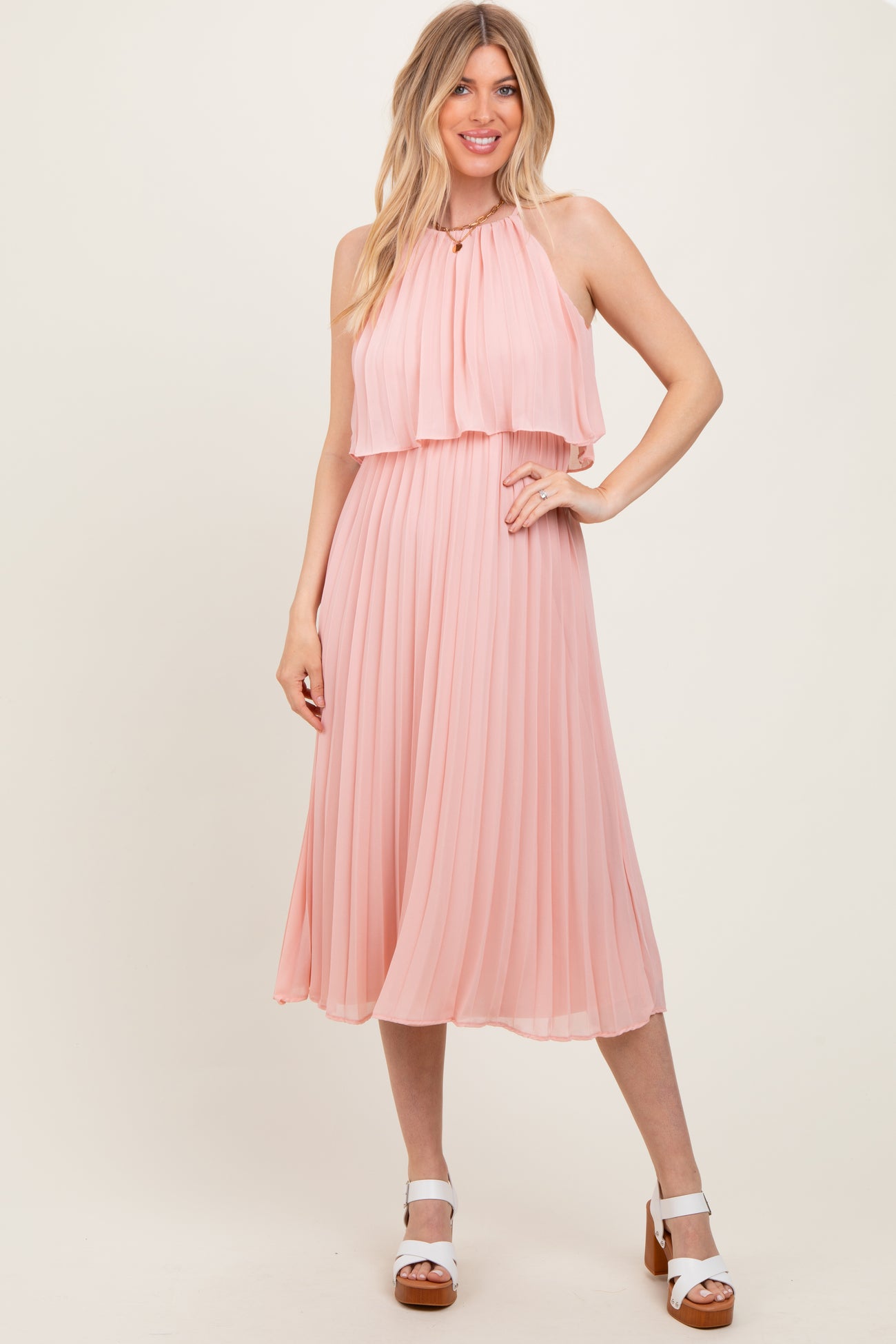 woman wearing a light pink pleated midi dress with a halter neck and layered top, standing in a neutral background