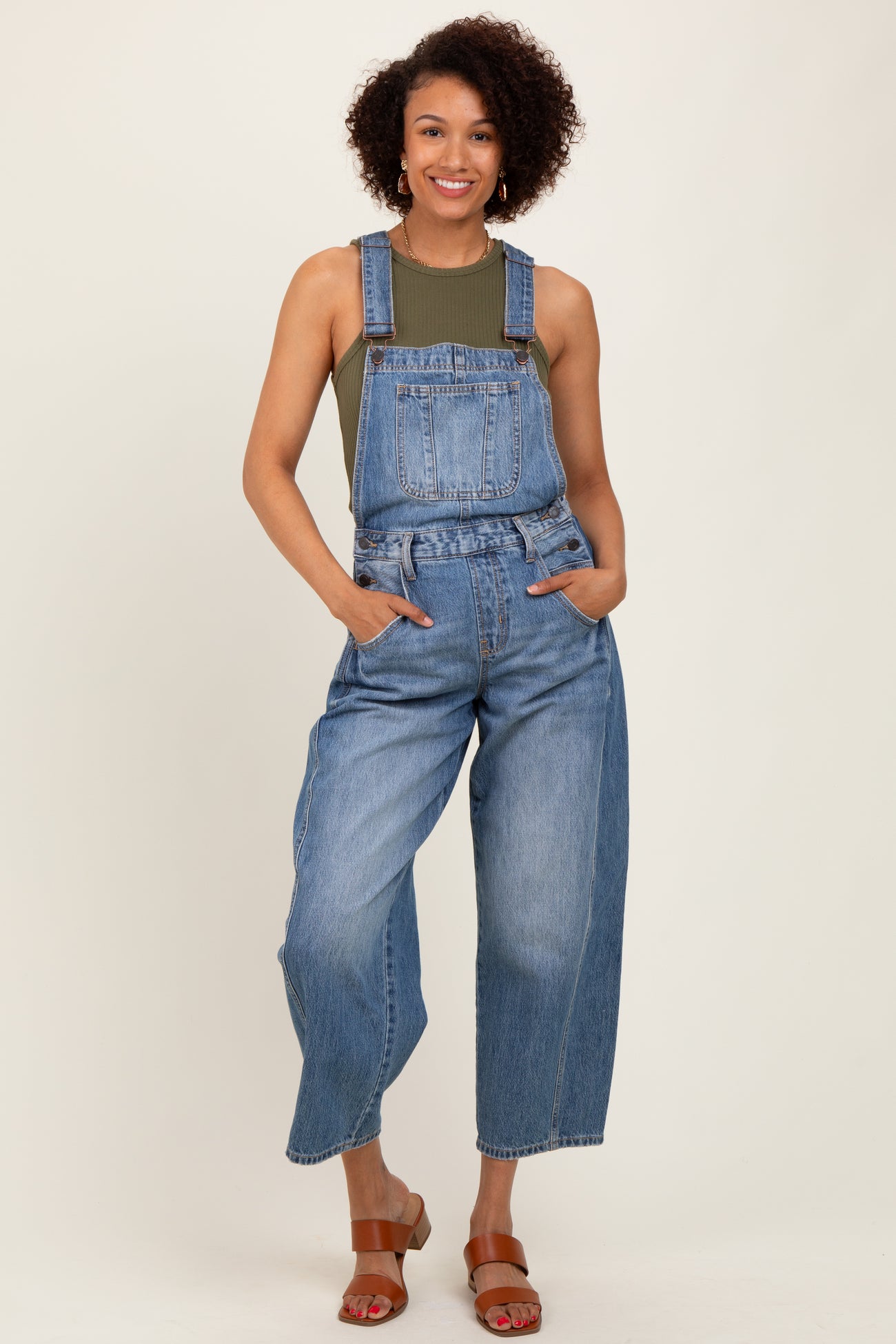 woman wearing a green sleeveless top and blue denim overalls with wide legs, standing with hands in pockets
