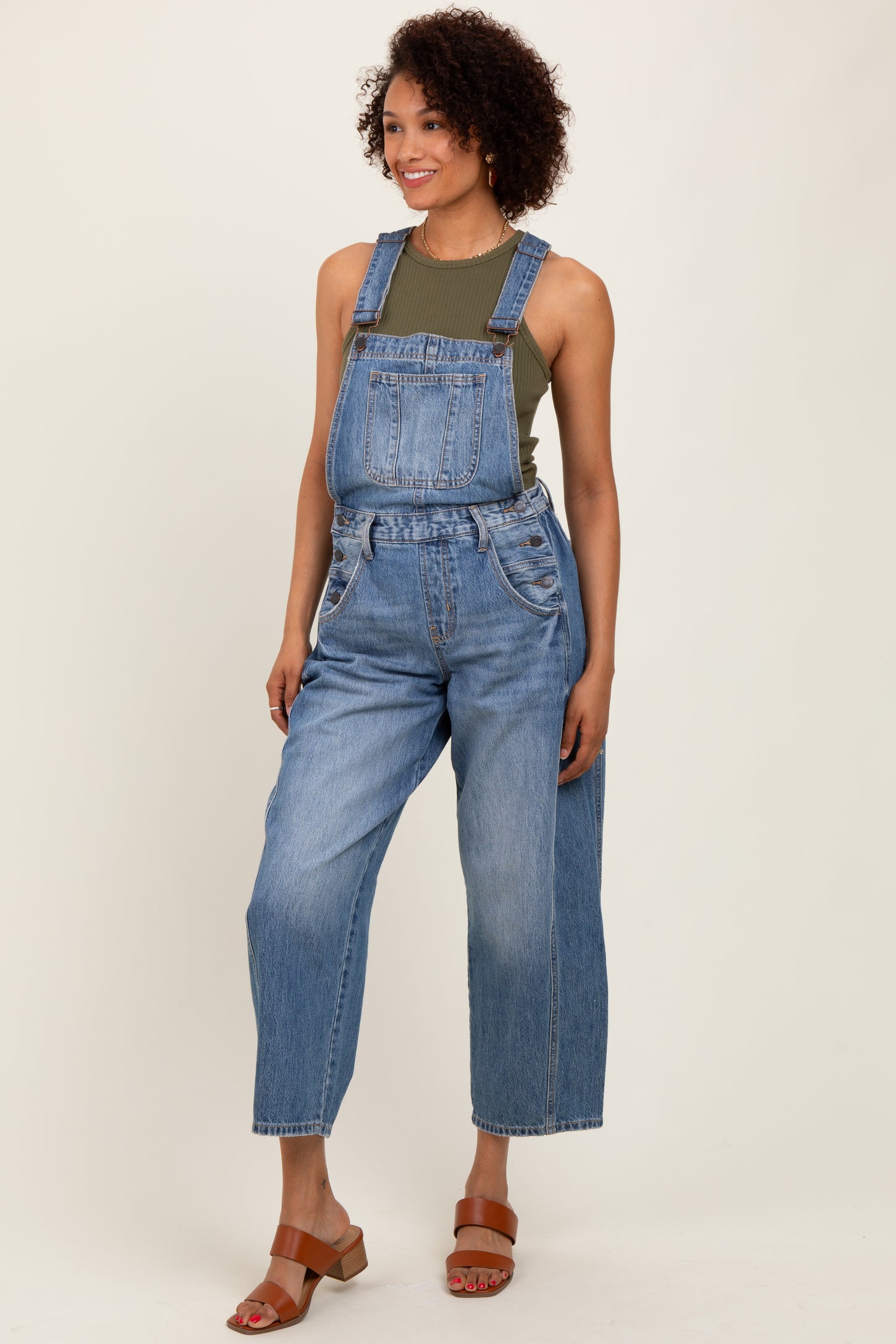 woman wearing light wash denim overalls with a green tank top underneath and brown sandals