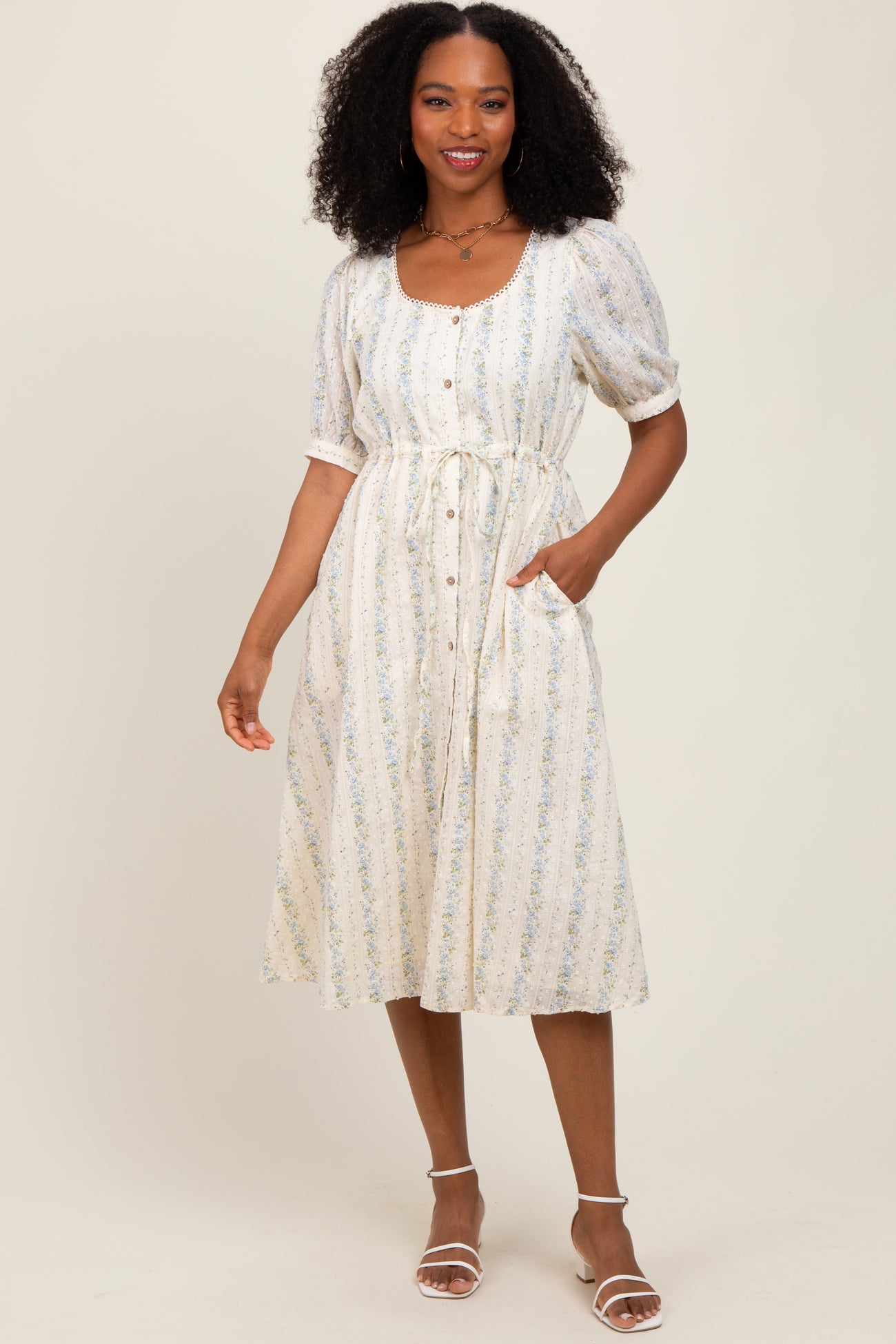 woman wearing a white floral midi dress with puff sleeves, button details, and a cinched waist, posing with hands in pockets
