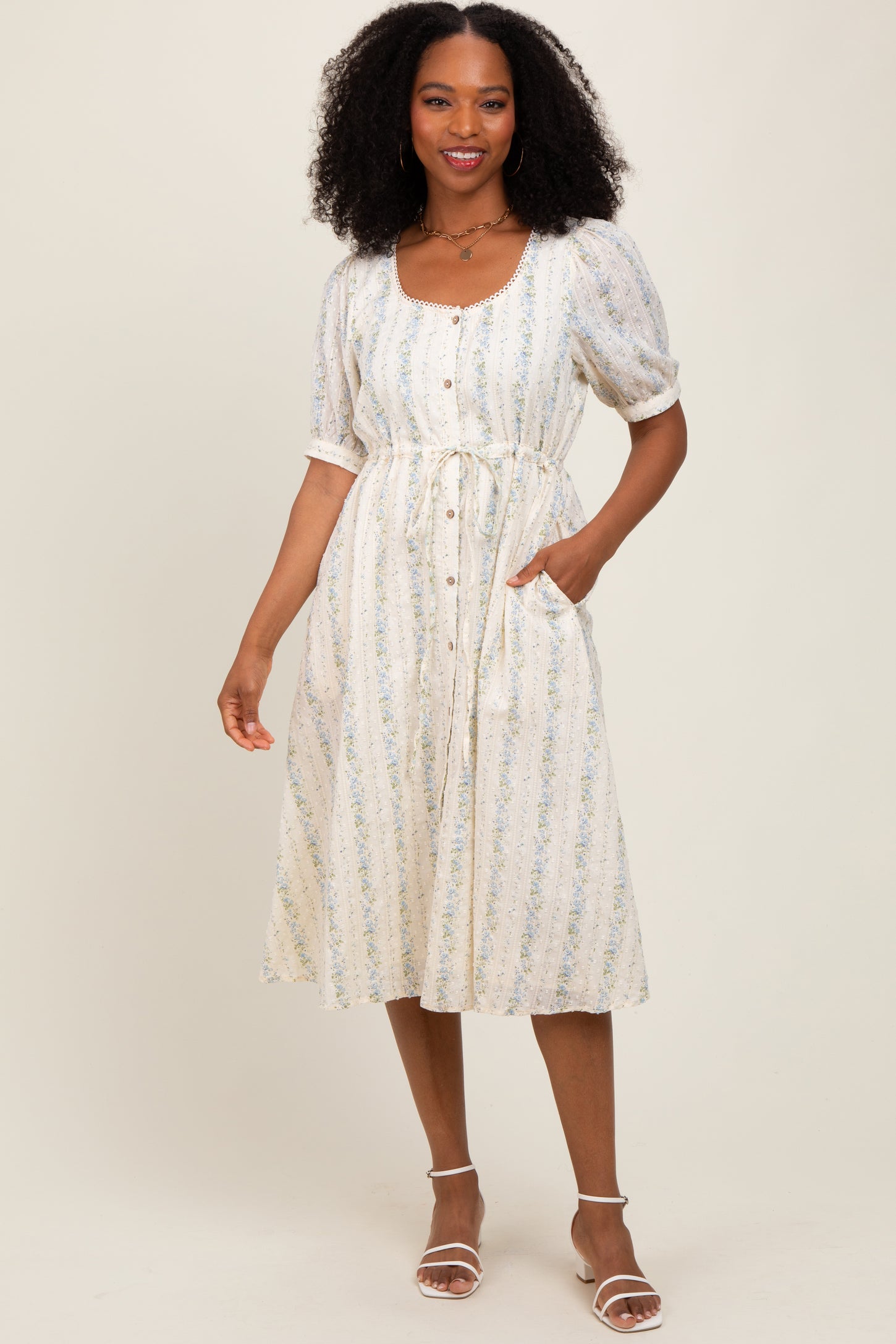 woman wearing a white floral midi dress with puff sleeves, button details, and a cinched waist, posing with hands in pockets
