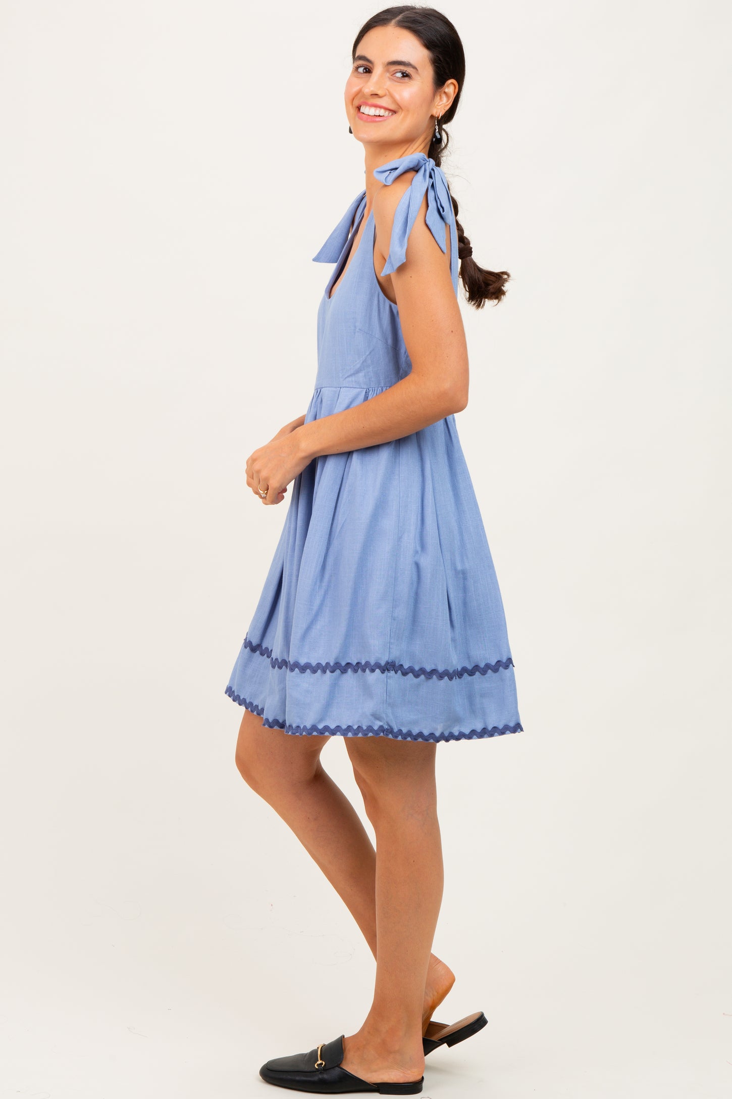 woman wearing a light blue sleeveless dress with ties on the shoulders and scalloped hem, smiling and standing sideways
