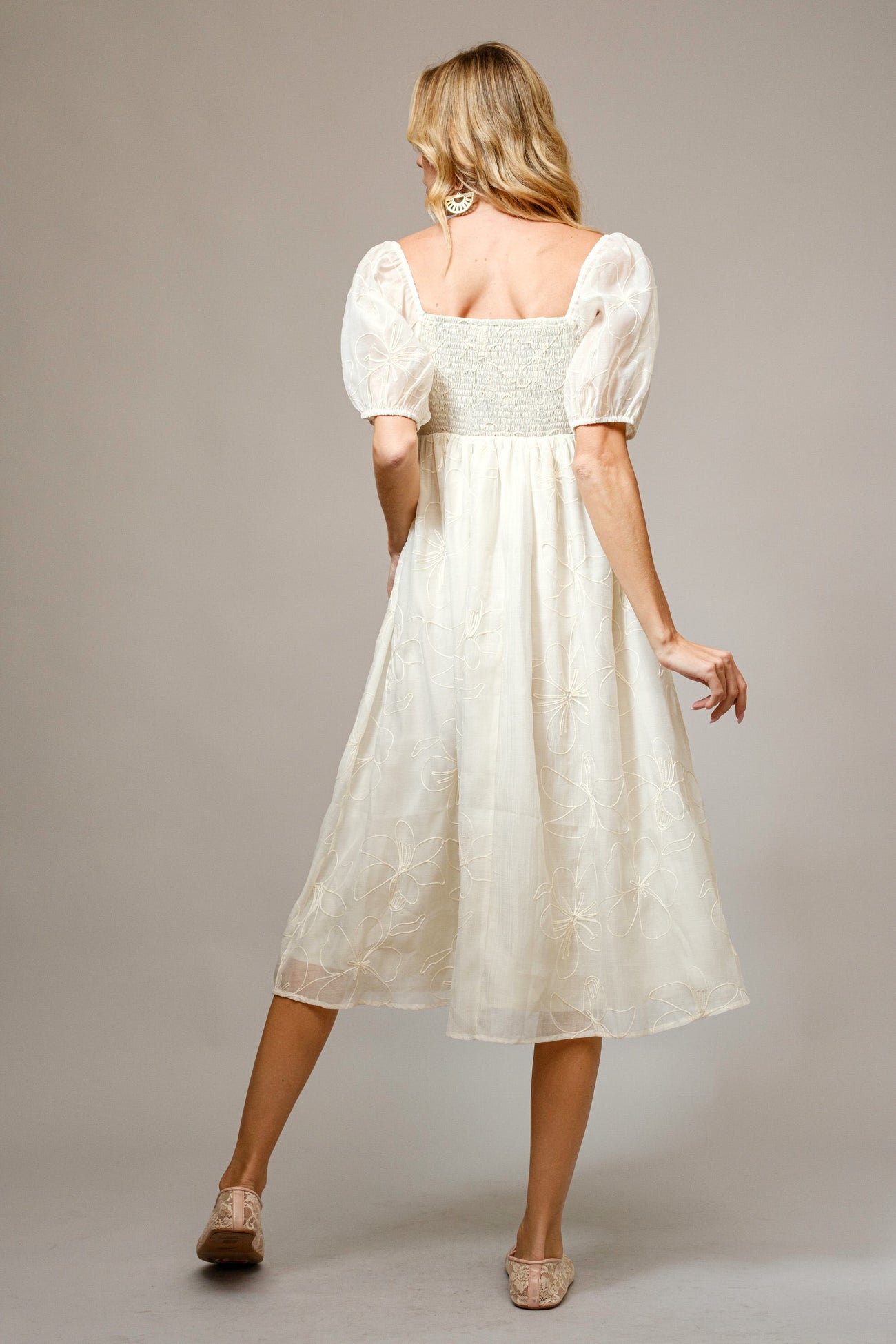 woman wearing a cream-colored midi dress with puff sleeves and a smocked bodice, standing with her back to the camera