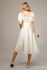 woman wearing a cream-colored midi dress with puff sleeves and a smocked bodice, standing with her back to the camera