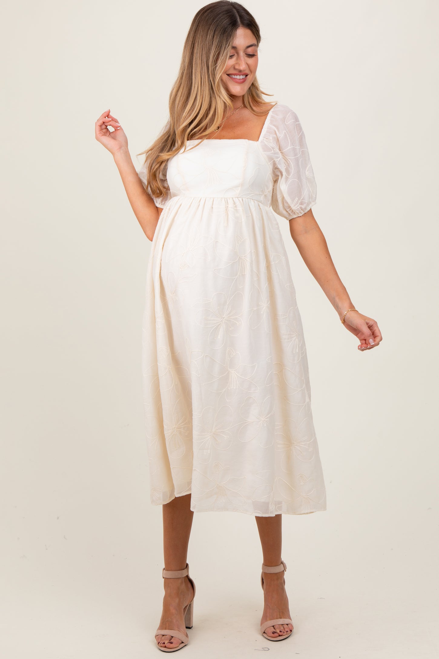 woman wearing a cream maternity dress with puff sleeves and floral embroidery, gently cradling her baby bump