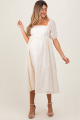 woman wearing a cream maternity dress with puff sleeves and floral embroidery, gently cradling her baby bump