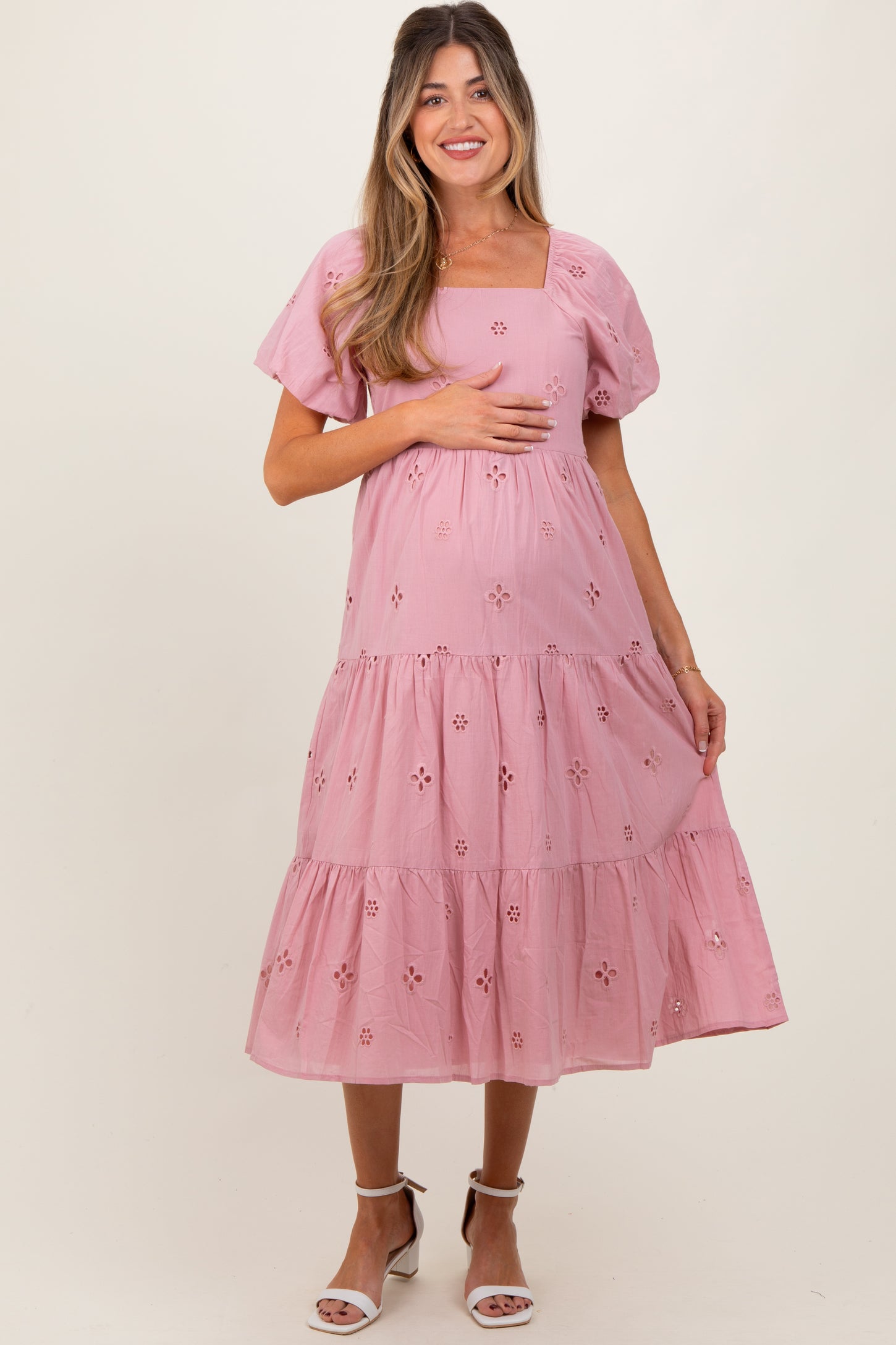 woman wearing a pink embroidered maternity dress with a square neckline and tiered skirt, holding her baby bump