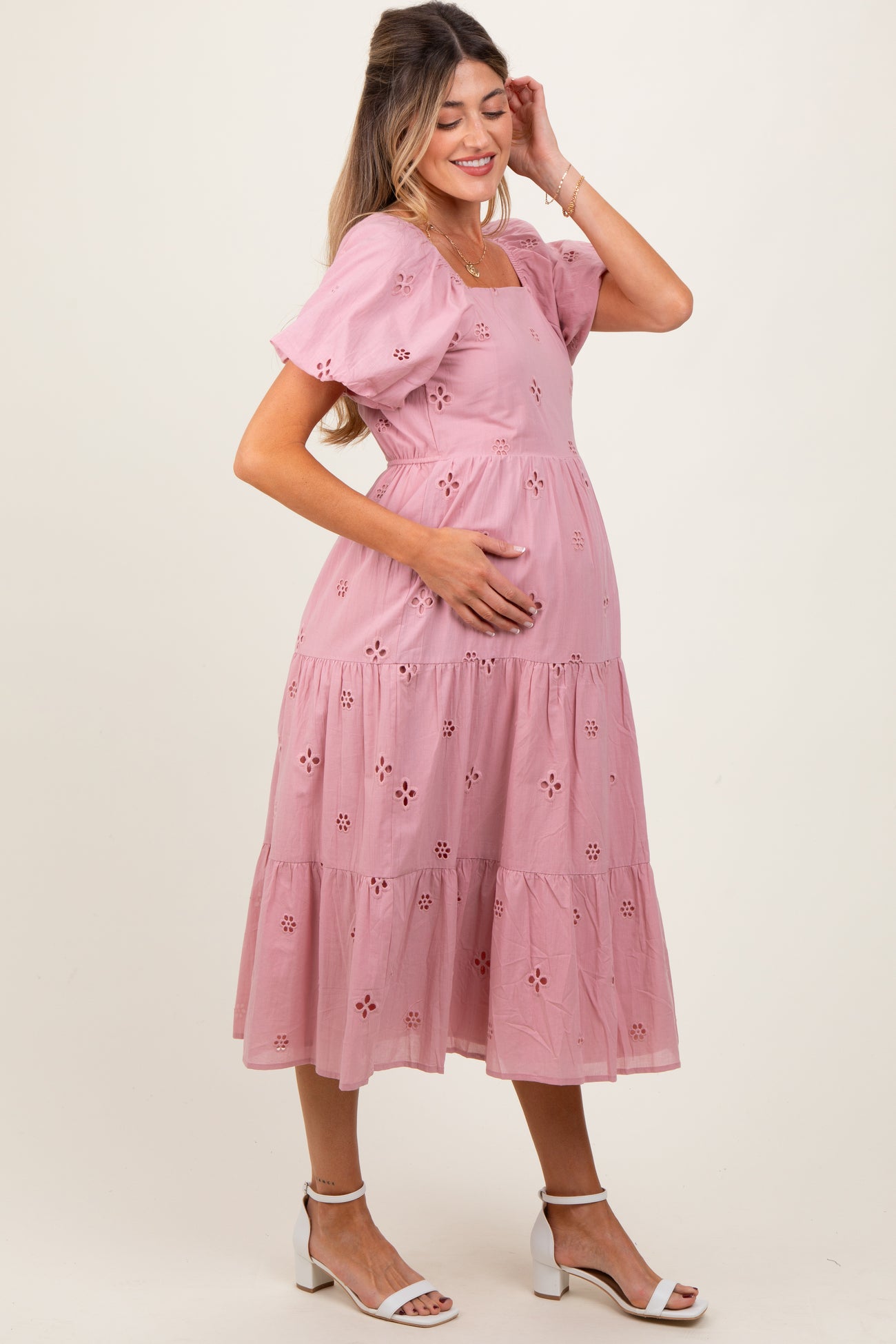 woman wearing a pink maternity dress with puff sleeves and eyelet details, gently cradling her baby bump