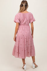 woman wearing a pink maternity dress with short puff sleeves and floral embroidery, showing a gathered waist and tiered skirt