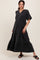 woman wearing a black tiered maternity dress with short sleeves and button details, standing in a relaxed pose