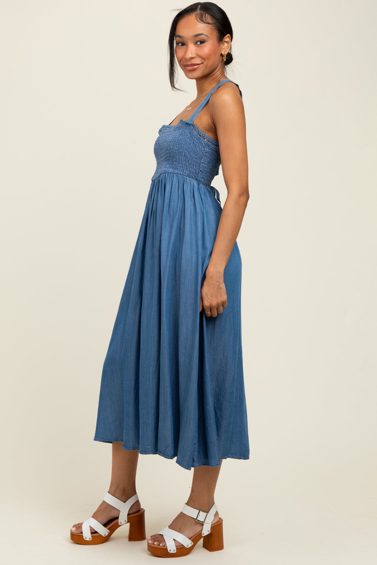 woman wearing a denim blue midi dress with smocked bodice and spaghetti straps, paired with white block heel sandals