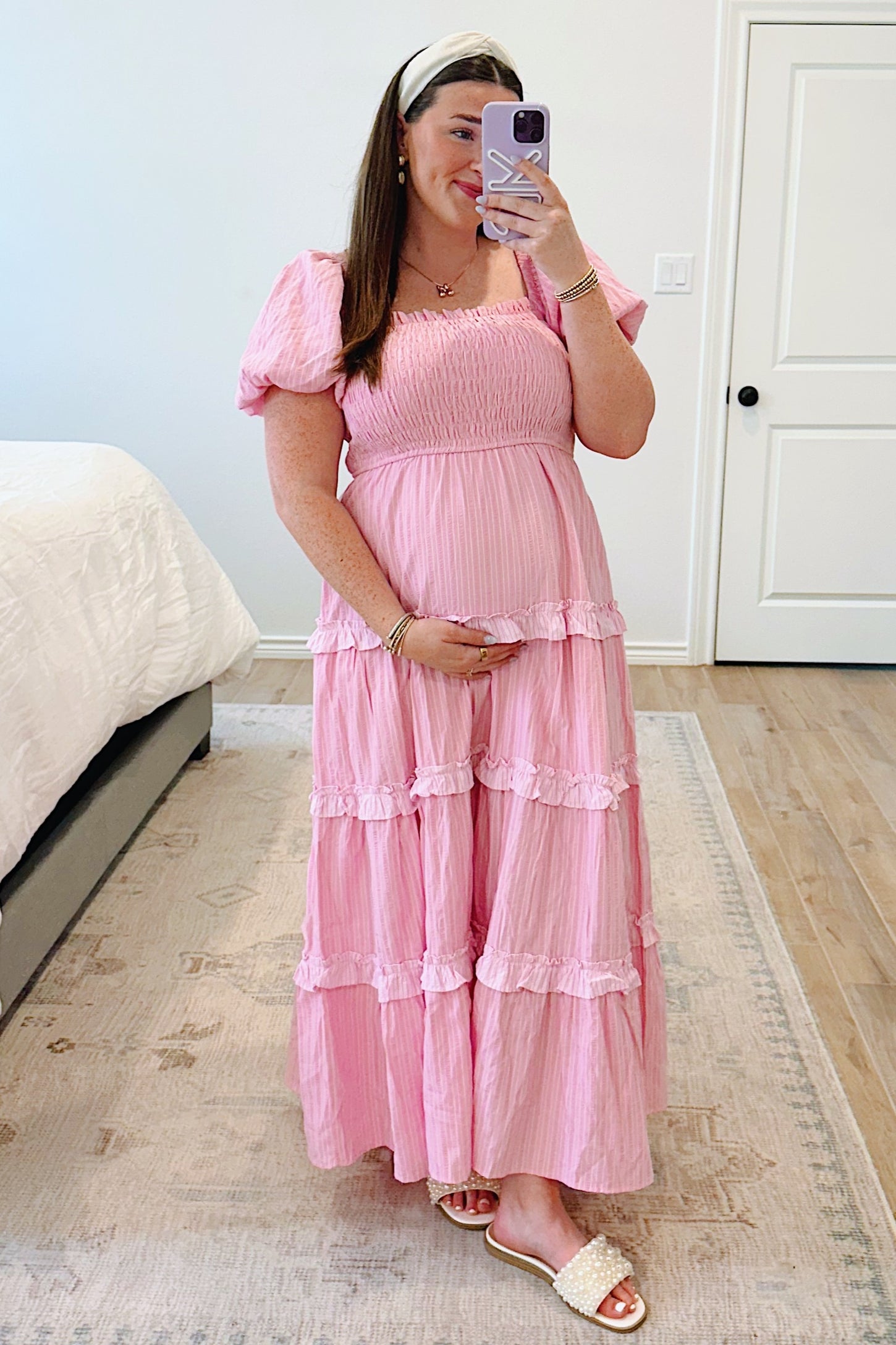 woman wearing a pink tiered maternity dress with puff sleeves and a baby bump visible, standing in a bedroom setting
