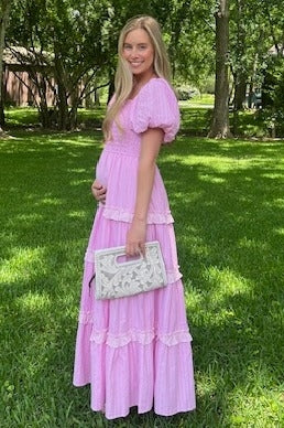 woman wearing a pink maternity maxi dress with puff sleeves and ruffled detailing, holding a clutch and showing her baby bump