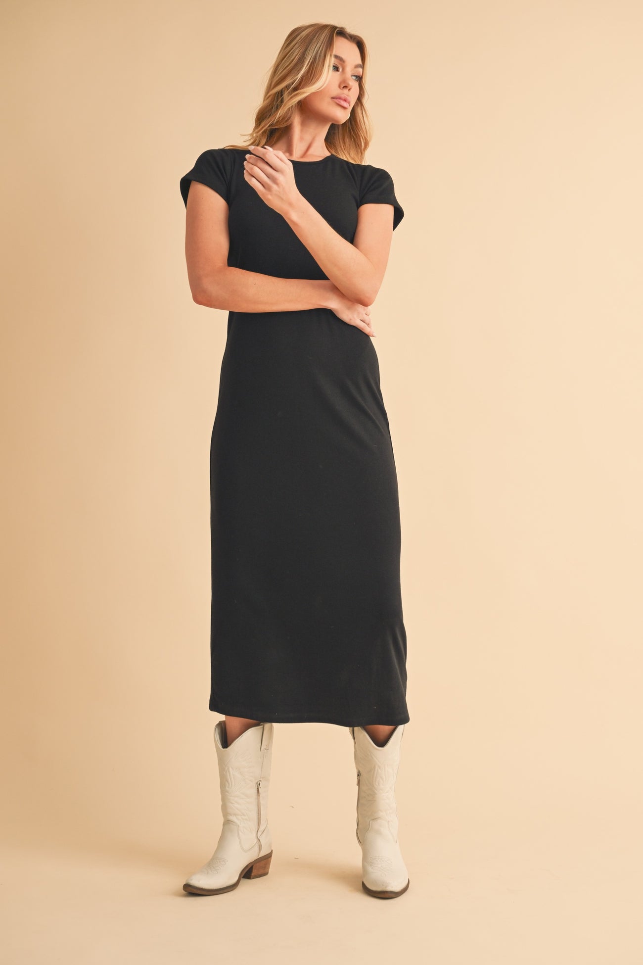 woman wearing a black cap sleeve midi dress and white cowboy boots, posing against a neutral background