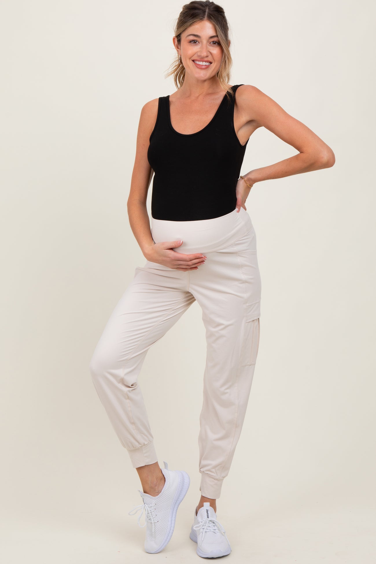 woman wearing a black maternity tank top and cream jogger pants, holding her baby bump