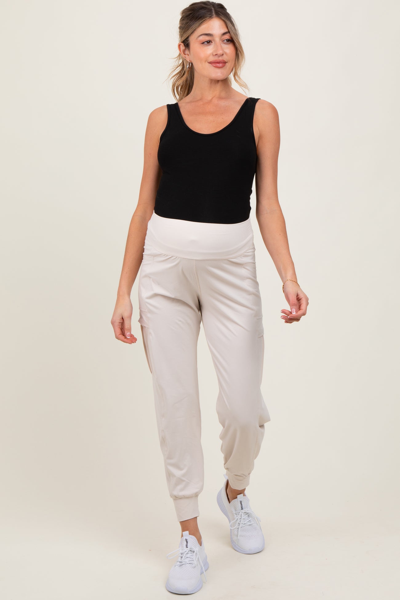 woman wearing a black tank top and cream maternity joggers with a full belly panel, styled with white sneakers