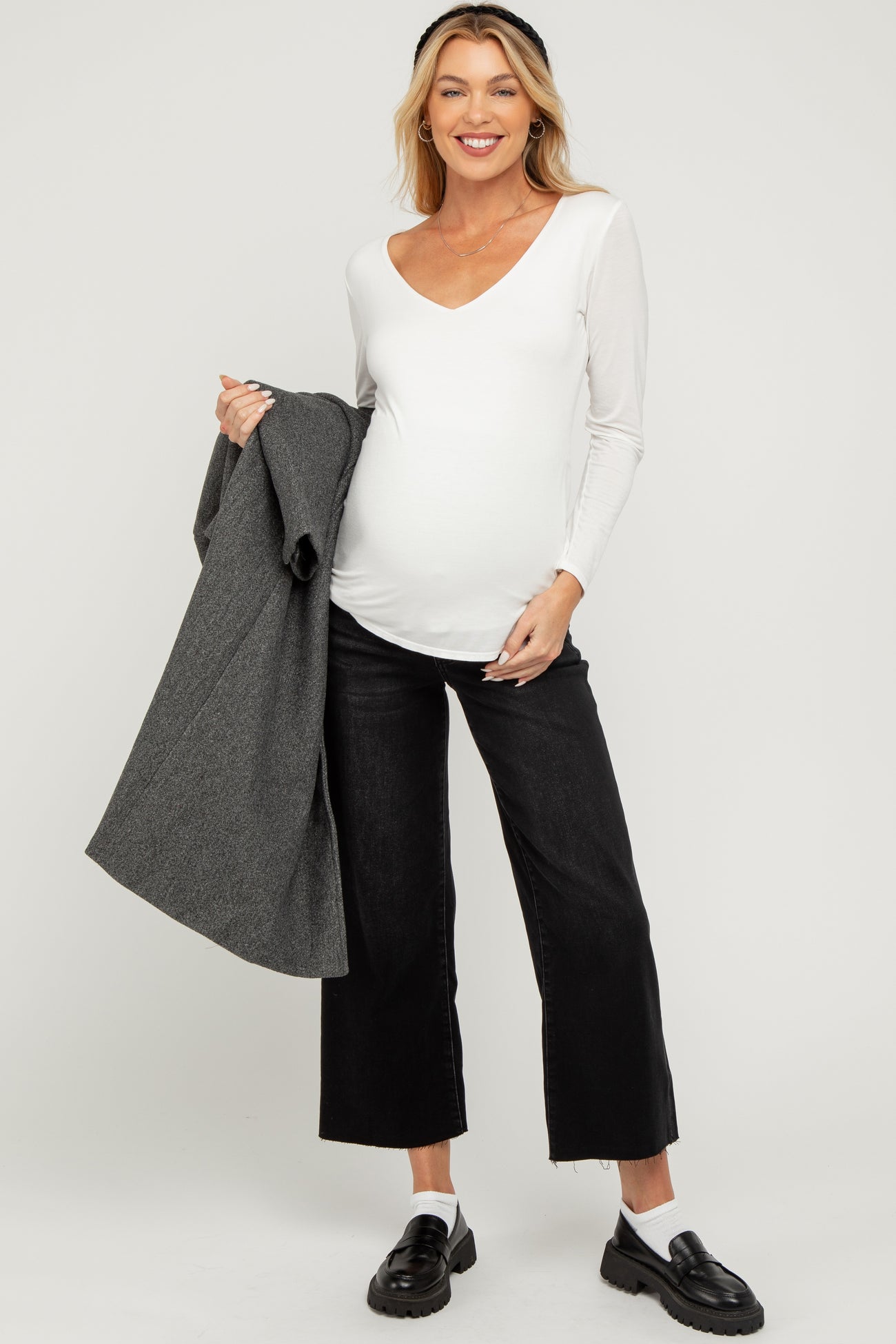 woman wearing a white long sleeve maternity top and black wide-leg jeans, holding a gray jacket, with a visible baby bump