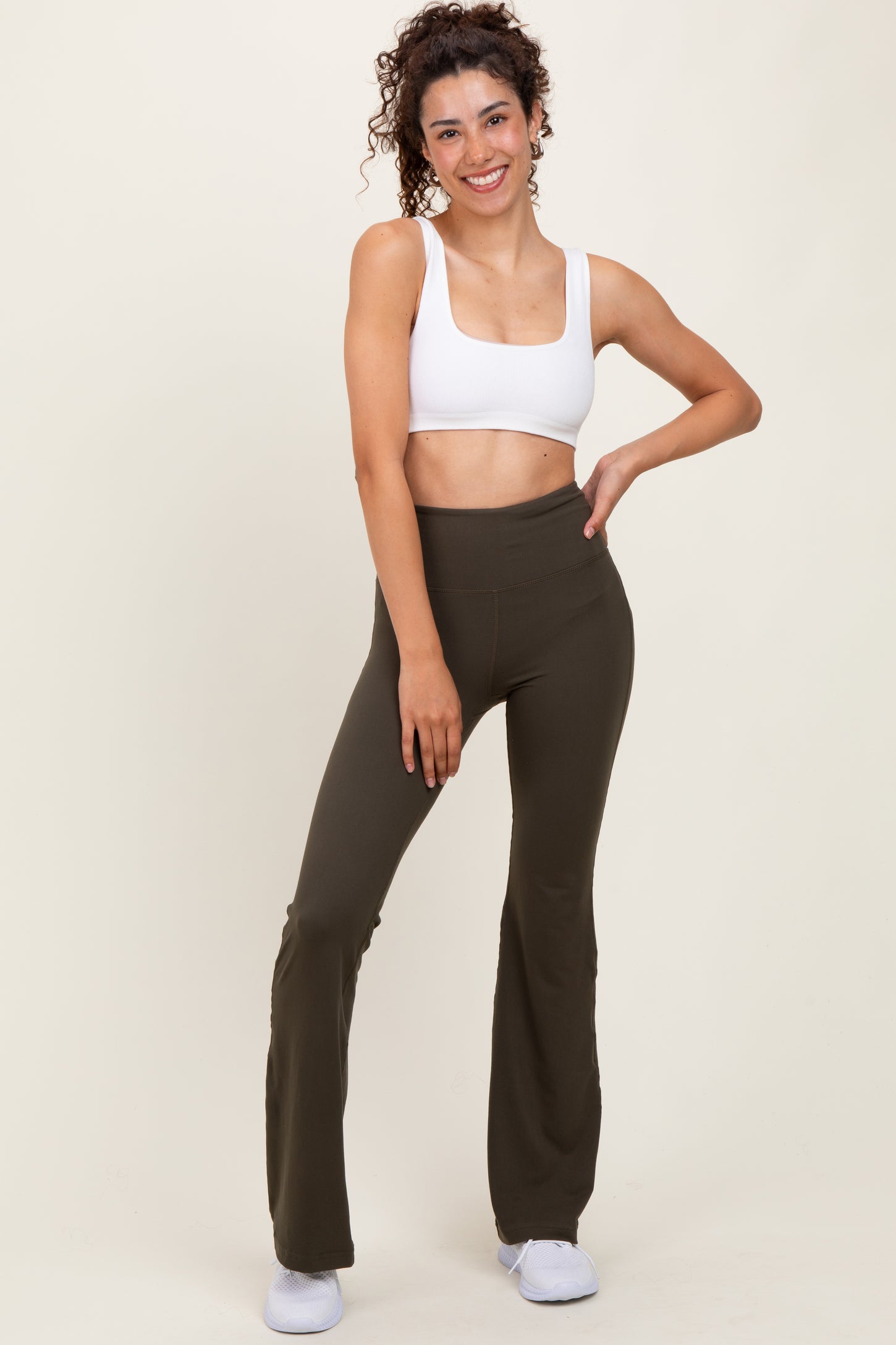 woman wearing a white crop top and olive green flare yoga pants, standing with a smile against a neutral background