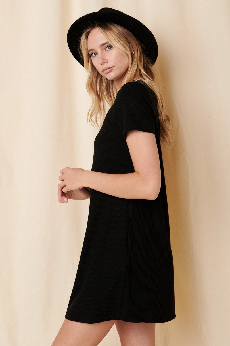 woman wearing a black short sleeve ribbed dress and a black hat, standing sideways against a light backdrop